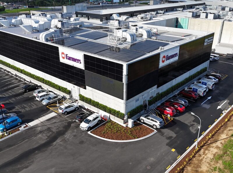 Aerial view of a large, two-story commercial building (Farmers store at Tauranga Crossing) featuring a solar array on its flat roof and some vertical solar panel mounting visible on the upper exterior wall, adjacent to the roofline.