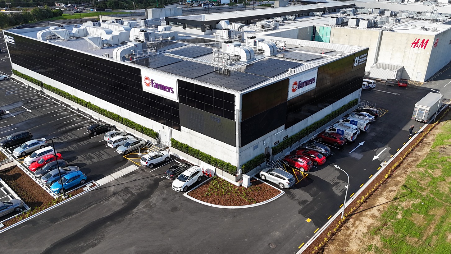 Aerial view of a large, two-story commercial building (Farmers store at Tauranga Crossing) featuring a solar array on its flat roof and some vertical solar panel mounting visible on the upper exterior wall, adjacent to the roofline.